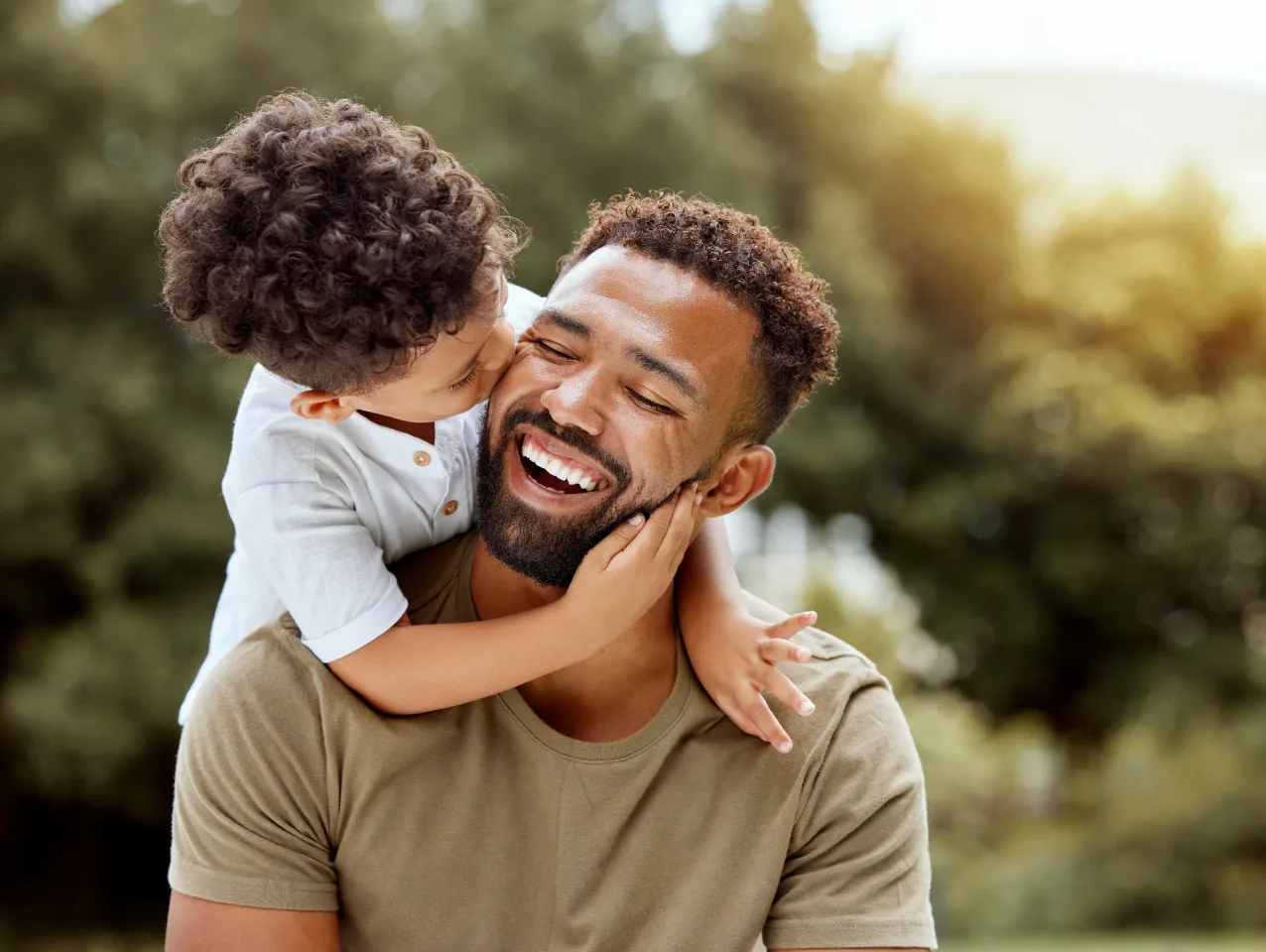 A dad laughing as his young son hugs and kisses him outdoors.